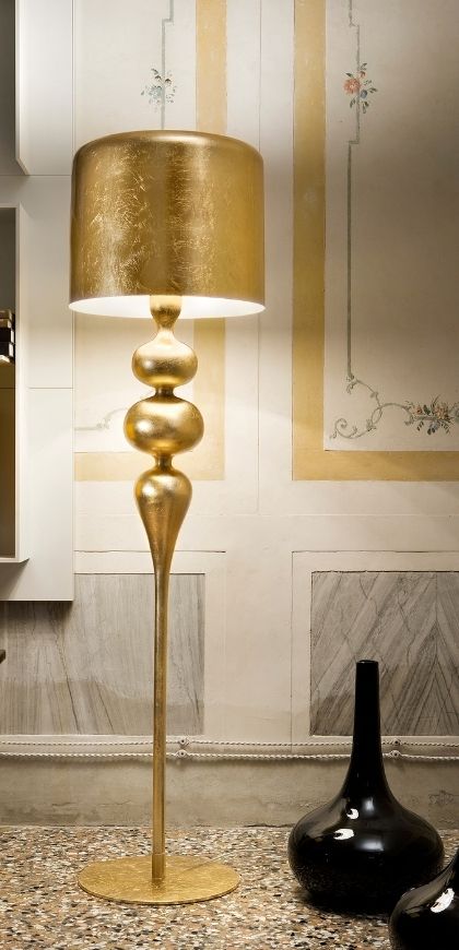 Goldene Stehleuchte Eva von Masiero mit Blattgold-Schirm und geschwungenem Schaft vor einer Wand mit gemalten Blumenranken
